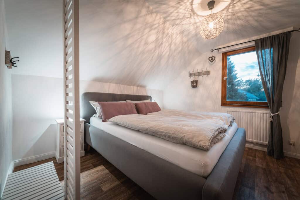 Ein gemütliches Schlafzimmer in einem Ferienhaus in der Eifel mit einem großen grauen Bett, weißer Bettwäsche, rosa Kissen, Holzboden und einem Fenster mit dunklen Vorhängen, das Tageslicht hereinlässt. Eine moderne Pendelleuchte wirft Schattenmuster an die Decke.