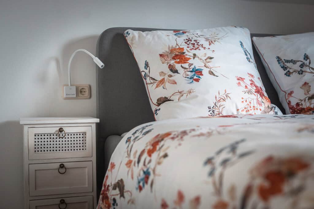 Ein ordentlich gemachtes Bett mit weißer Bettwäsche mit Blumenmuster steht neben einem kleinen weißen Nachttisch. Dieses Ferienhaus Monschau bietet mit seiner flexiblen Leselampe über der Steckdose perfekten Komfort zum Entspannen nach einem Tag in Monschau.