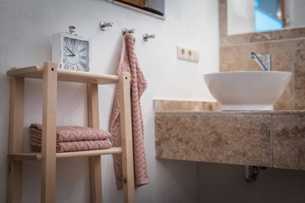Ein Badezimmer im Ferienhaus Monschau verfügt über ein Holzregal mit gefalteten Handtüchern und einer weißen Uhr, ein an der Wand hängendes Handtuch und eine Marmortheke mit einem runden weißen Waschbecken – perfekt für Ihren Urlaub in Monschau.