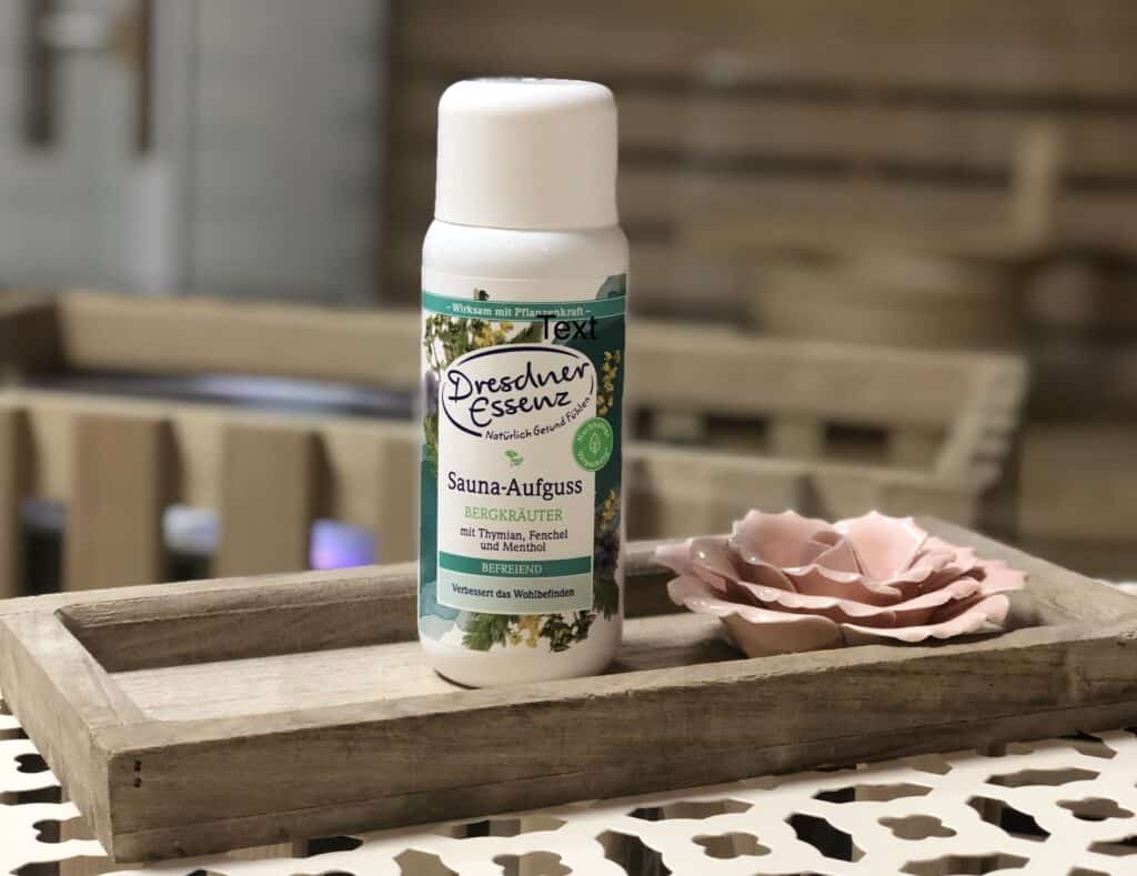 Eine Flasche Dresdner Essenz Saunaaufguss steht auf einem Holztablett neben einer rosa Keramikblume, perfekt zum Entspannen in Ihrem Ferienhaus Sauna Eifel, mit einem unscharfen Hintergrund mit gemütlichem Holzdekor.