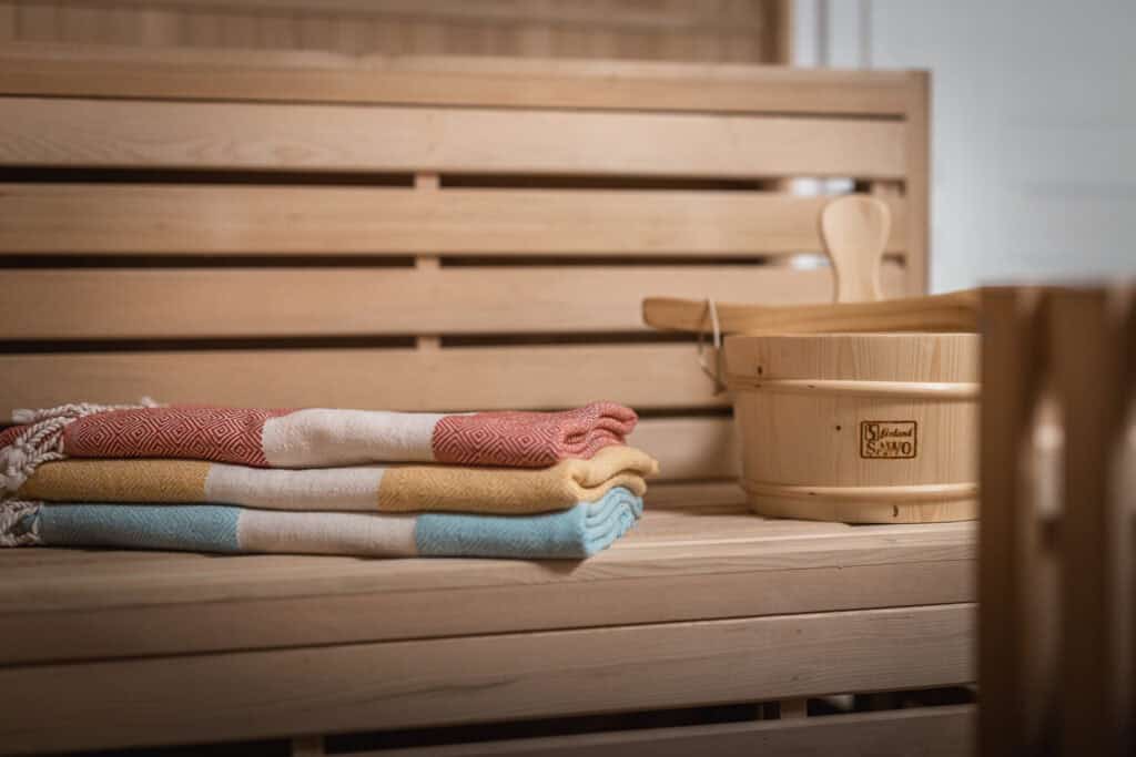Eine hölzerne Saunabank in Ihrem Ferienhaus in Monschau mit einem Stapel aus drei bunten Handtüchern und einem hölzernen Saunakübel mit Schöpfkelle daneben – perfekt zum Entspannen während Ihres Familienurlaubs in der Eifel.