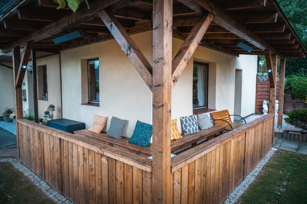Eine gemütliche Holzterrasse mit einer Bank mit bunt gemusterten Kissen, die an ein cremefarbenes Monschauer Ferienhaus angeschlossen ist. Die überdachte Terrasse, umgeben von einem Holzzaun, ist ideal für einen Familienurlaub in der Eifel, umgeben von üppigem Grün.