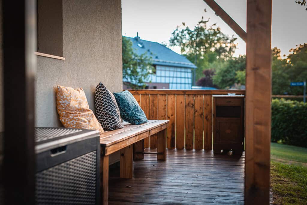 Eine Holzveranda mit einer Bank mit bunten Kissen überblickt einen grünen Hof, perfekt für einen Familienurlaub in der Eifel, mit einem kleinen Holzschrank und benachbarten Häusern, die im Hintergrund bei Sonnenuntergang sichtbar sind.
