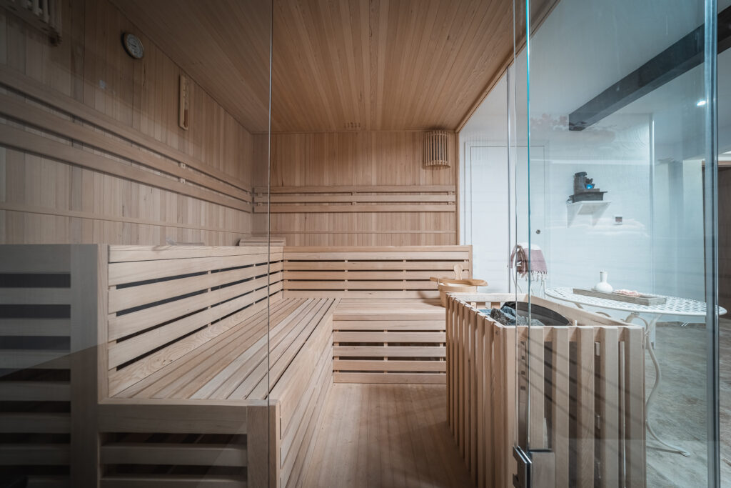 Eine moderne Holzsauna mit Glaswänden, Holzbänken, Uhr und Ofen in der Ecke, draußen ein Regal mit Handtüchern und Deko – perfekt zum Entspannen in Ihrem Ferienhaus Monschau oder für ein Ferienhaus Sauna Eifel-Erlebnis.