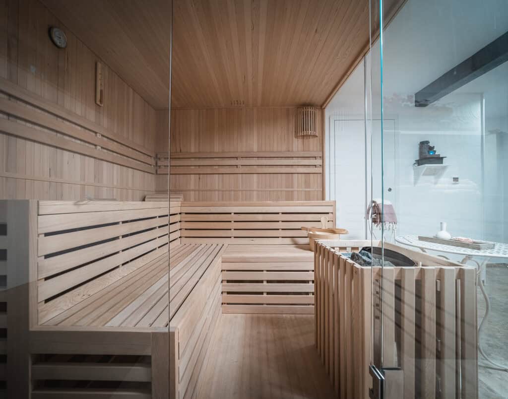 Eine moderne Holzsauna mit Glaswänden, Holzbänken, Uhr und Ofen in der Ecke, draußen ein Regal mit Handtüchern und Deko – perfekt zum Entspannen in Ihrem Ferienhaus Monschau oder für ein Ferienhaus Sauna Eifel-Erlebnis.