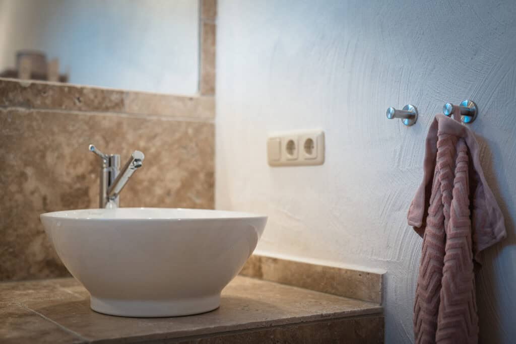 Ein modernes Badezimmerwaschbecken mit einem runden weißen Becken auf einer braunen Steinplatte, ideal für einen Familienurlaub in der Eifel – zur Ausstattung gehören ein Chromhahn, ein großer Spiegel, Steckdosen und ein rosa Handtuch, das an der Wand hängt.