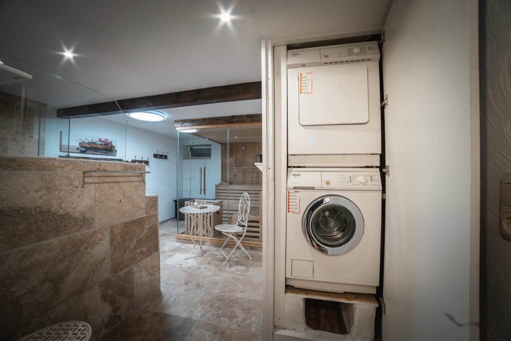 Ein moderner Waschraum mit Waschmaschine und Trockner befindet sich neben einer verglasten Sauna – ideal zum Entspannen nach einem Eifel-Ausflug mit Kindern oder einem Eifel-Wochenende in Ihrem Ferienhaus Sauna Eifel. Steinfliesenboden, ein kleiner Tisch und Einbauleuchten runden den Raum ab.