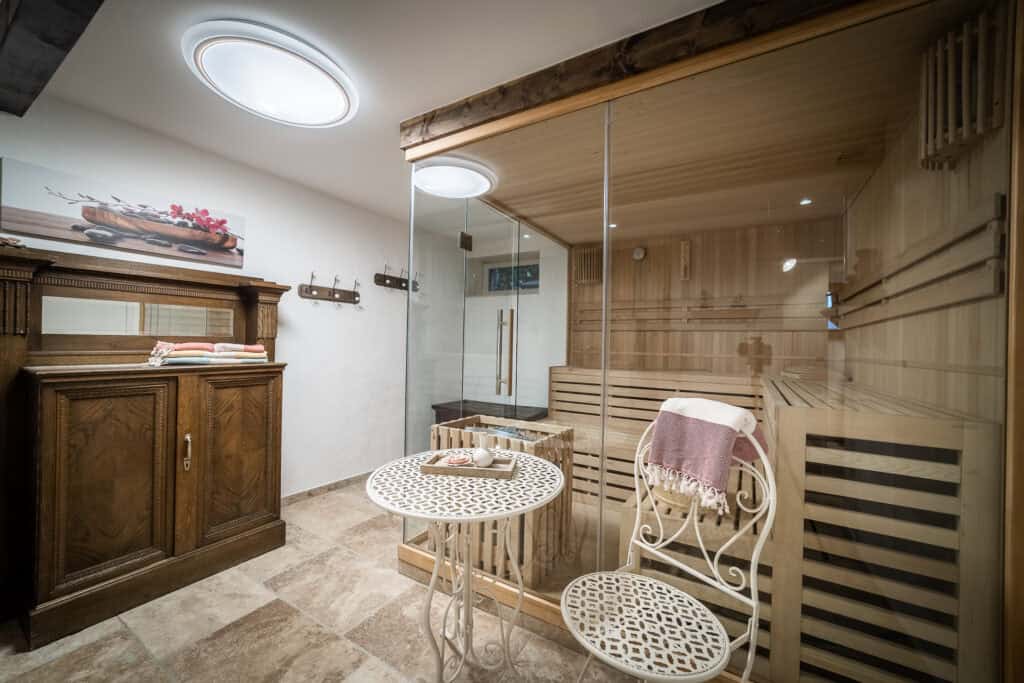 Ein gemütlicher Wellnessraum in einem Monschauer Ferienhaus mit einer Holzsauna mit Glaswänden, einem kleinen runden Tisch mit zwei verzierten Metallstühlen, Handtüchern auf einem Holzschrank und beigefarbenem Fliesenboden. Die sanfte Beleuchtung trägt zur einladenden Atmosphäre bei.
