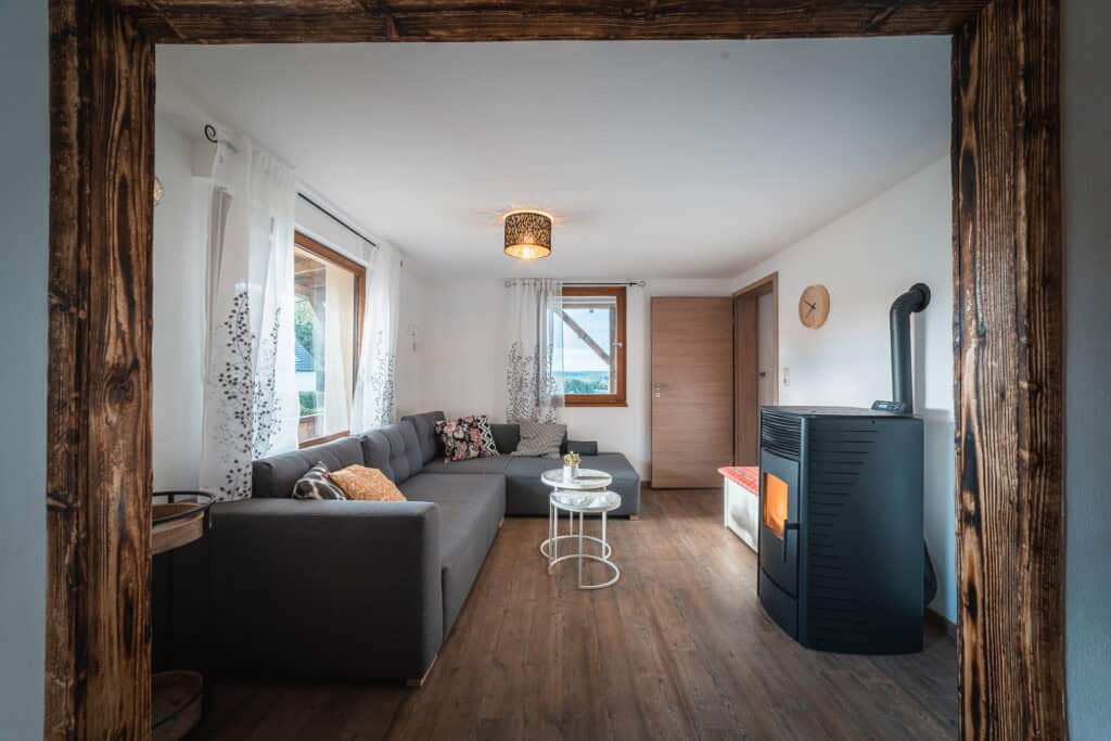 Gemütliches Wohnzimmer mit grauem Ecksofa, runden Couchtischen und großen Fenstern mit weißen Vorhängen – ideal für ein Eifelwochenende oder einen Aufenthalt im Ferienhaus in Monschau. Warme Beleuchtung und ein Holzofen verleihen den rustikalen Holzböden Charme und eine einladende Atmosphäre.