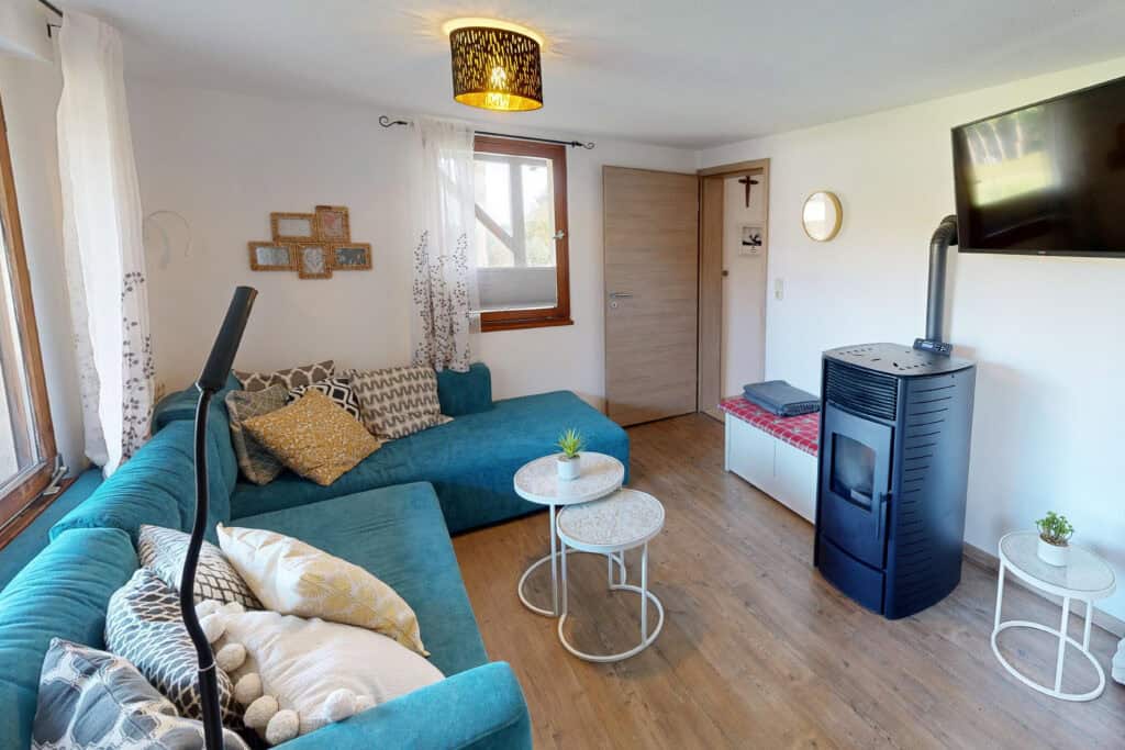 Ein gemütliches Wohnzimmer in einem Ferienhaus Eifel mit einem blaugrünen Sofa, gemusterten Kissen, zwei runden weißen Tischen, einem Holzofen, einem Flachbildfernseher an der Wand und großen Fenstern, die den Raum in natürliches Licht tauchen.