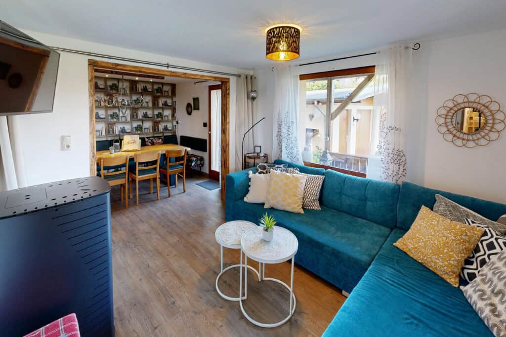 Ein gemütliches Wohnzimmer mit einem blaugrünen L-förmigen Sofa, verschiedenen Kissen, zwei runden weißen Couchtischen und großen Fenstern mit transparenten Vorhängen – perfekt für einen erholsamen Urlaub in Monschau in Ihrem charmanten Ferienhaus Eifel.