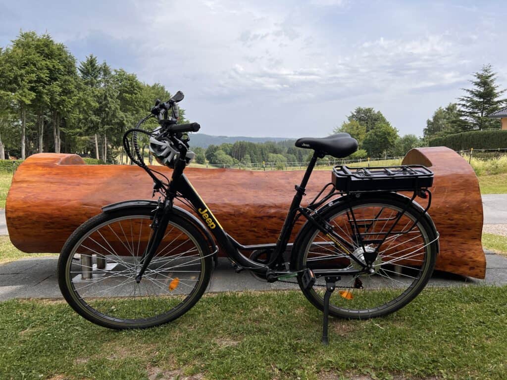 Ein schwarzes Jeep-Fahrrad steht auf einem gepflasterten Weg vor einer großen, polierten Holzbank in der Nähe von Bäumen und Grasfeldern, perfekt für Erkundungstouren während des Aufenthalts im Ferienhaus Monschau unter einem teilweise bewölkten Himmel.