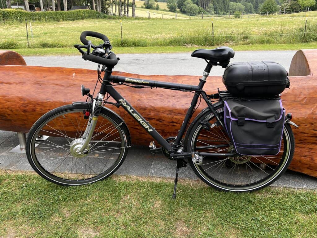 Ein schwarzes Fahrrad mit einer Heckbox und einer Seitentasche steht auf einem Weg in der Nähe einer langen Holzbank – perfekt für alle, die in einem Ferienhaus Monschau übernachten und die ruhigen Wiesen und Bäume in der Eifel genießen möchten.