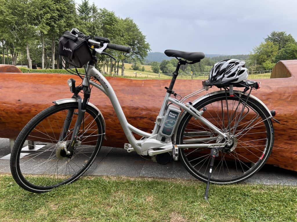 Ein silbernes Elektrofahrrad mit schwarzem Sattel, Vordertasche und Helm auf dem Gepäckträger steht auf dem Rasen neben einem großen Holzstamm – perfekt, um während eines unvergesslichen Urlaubs in Monschau die Hügel und Bäume in der Nähe Ihres Ferienhauses Eifel zu erkunden.